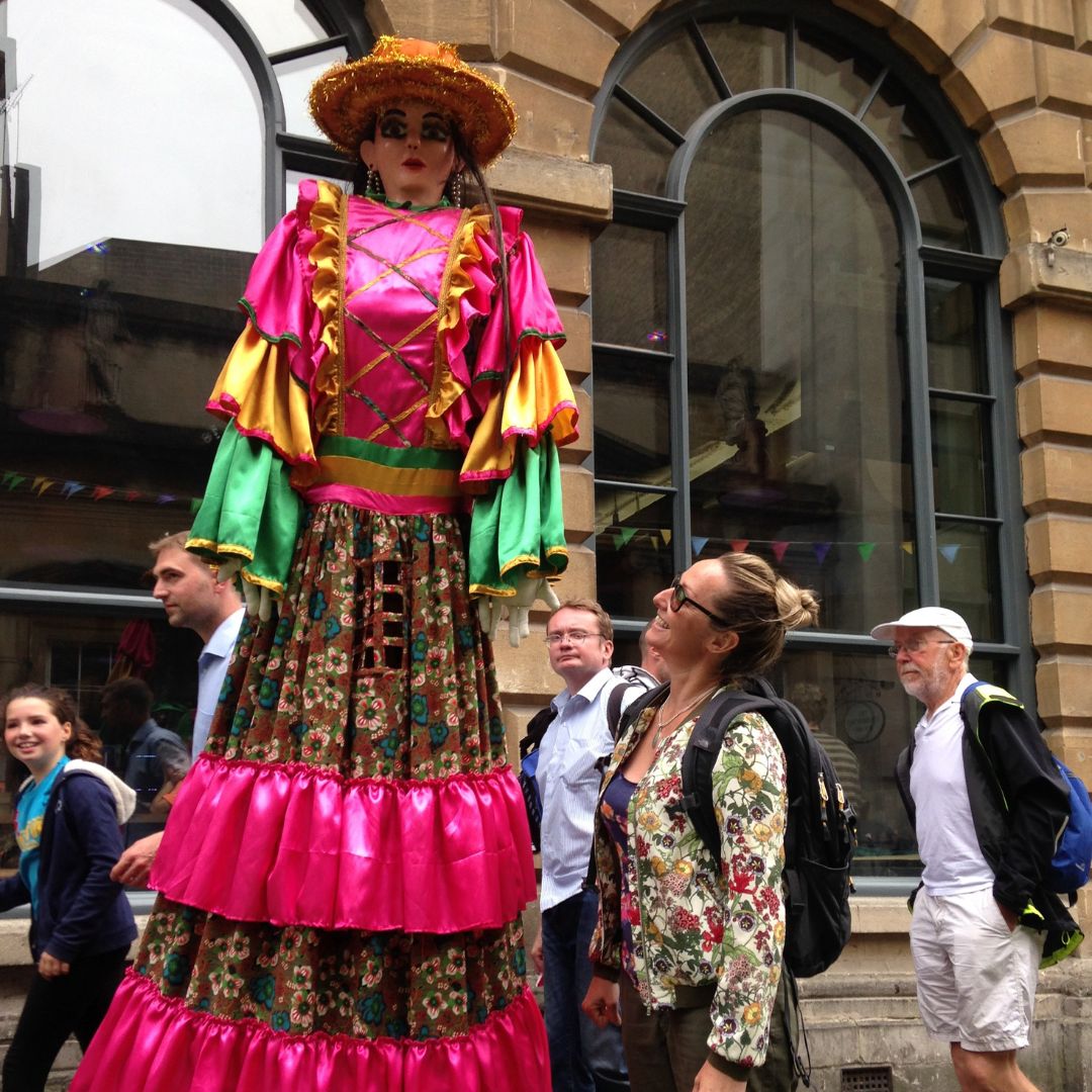 Kelly in the street looking up at a street performer walking on stilts. The performer is wearing a pink dress and has a mask on